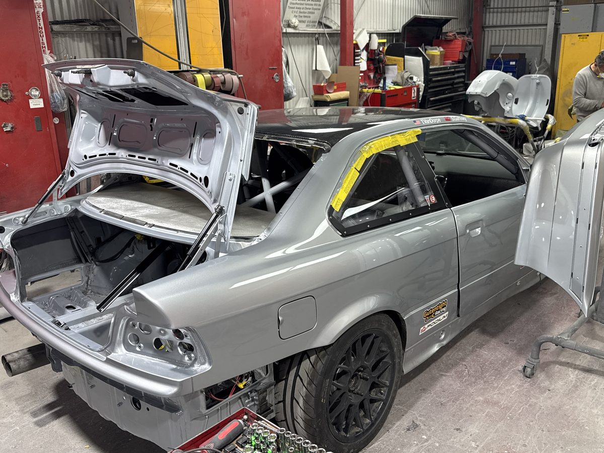 A technician sanding a panel in the workshop beside a BMW coupe with its trunk lid and hood removed
