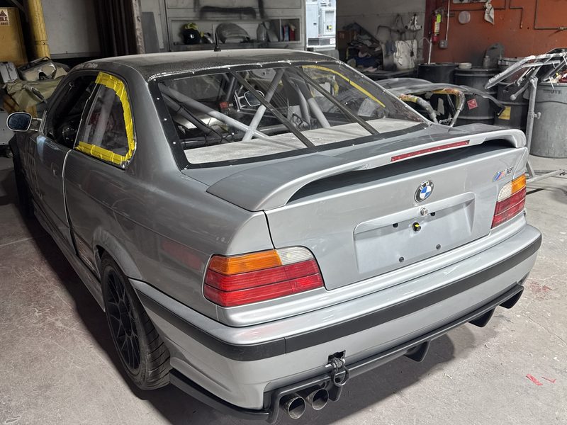 A silver BMW M3 coupe in the workshop with its trunk lid raised, a roll cage fitted and body panels on a stand alongside.