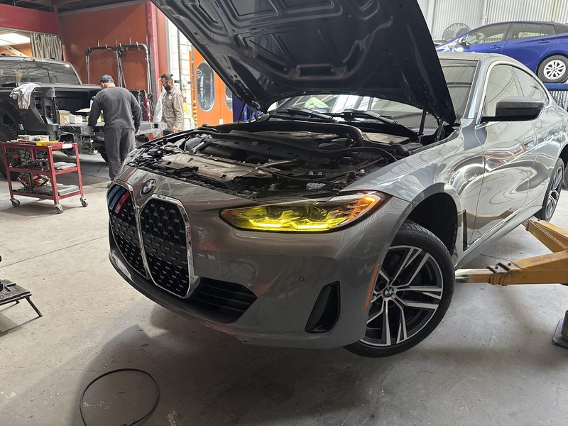 A grey BMW coupe in the workshop, hood raised, with a technician working at a bench in the background.