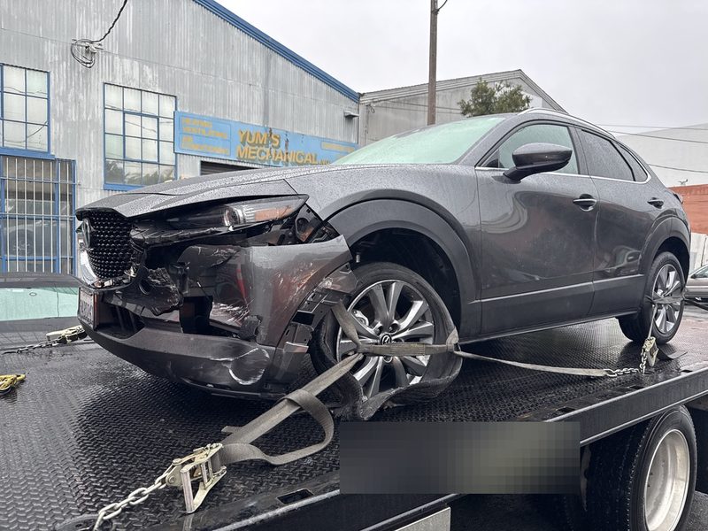 A grey crossover with heavy front-end damage strapped to the deck of a flatbed tow truck outside the shop.