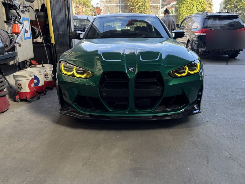 A BMW M4 finished in dark green with yellow-tinted headlights, parked at the front of the shop.