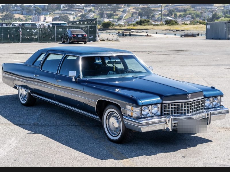 A dark blue 1970s Cadillac Fleetwood in gloss finish, photographed side on against an open lot.